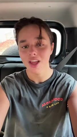 A young person with short, dark hair and a septum nose piercing is the central subject of the video. They wear a dark gray t-shirt with rolled-up sleeves, which features red and orange text reading "ALOHAMOOD A WAY OF LIFE."

The scene is set inside a vehicle, likely a car or jeep, viewed from the back seat. Dark upholstered seats are visible, and a window in the background shows a blurred exterior suggesting a dry, possibly dusty environment with reddish-brown ground and some sparse vegetation.

The person is actively singing along to music, lip-syncing with enthusiastic and expressive facial movements. They nod and move their head and shoulders in rhythm with the song. Their expressions convey joy, happiness, and engagement with the music, often smiling widely and playfully closing their eyes. The lighting indicates it is daytime, and the overall mood is cheerful and energetic.