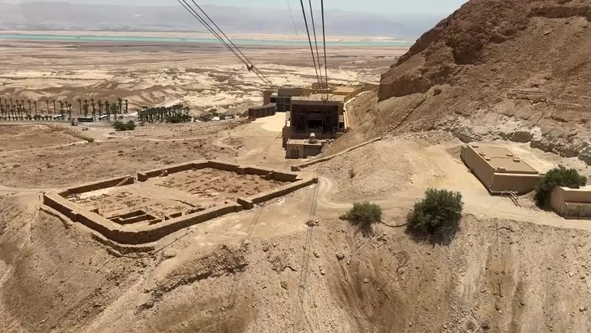 This video shows a panoramic view of an archaeological site on a rocky hill. The foreground features the excavated remains of stone structures, appearing as foundations and low walls. In the mid-ground, a cable car station is visible, with its supporting cables extending upwards towards the top of the hill. A cable car is suspended between the station and another point, suggesting it is in operation or paused.

The environment is arid and desert-like, with sparse vegetation, including a few small bushes and trees. In the background, a vast, dry landscape stretches out, leading to a body of water, possibly the Dead Sea, with distant mountains visible on the horizon. The sky is clear and bright, indicating a sunny day. There are no people clearly visible in the frame. The overall impression is one of ancient ruins set against a dramatic desert backdrop.