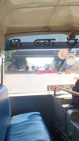 The camera looks out the front of a bus as it passes through a toll booth and enters an open highway.  The bus seat is a bright blue. The toll booth is clearly marked with red and white stripes, as are the many traffic signs lining the highway. A truck drives ahead, moving into the distance. Several other vehicles are visible far ahead as well.