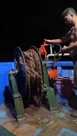 A man is operating a winch with a large, rusty chain coiled on it. He is standing to the right of the winch and appears to be controlling it with a lever. The scene is dark, suggesting it is nighttime, and the winch is mounted on a boat. The chain is being wound onto the winch.