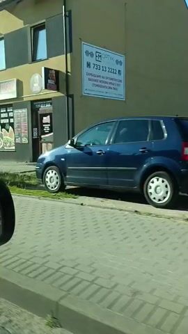 The video appears to be captured from a moving vehicle, likely a car, as evidenced by the side mirror visible in the bottom left corner of the frame and the changing scenery. The footage begins by showing a commercial building with a sign that reads "MAGIC KEBAB". The storefront displays images of food, suggesting it is a restaurant. The camera then pans to reveal an outdoor scene with a peculiar art installation on a lawn, featuring what looks like a giant blue haystack with a red cone on top. Further down the road, another building is visible, possibly a house with a fence. The video continues to show more buildings and an intersection with a traffic mirror. The environment seems to be a mix of commercial and residential areas under a clear blue sky, indicating daytime. There are no people visible in the video.