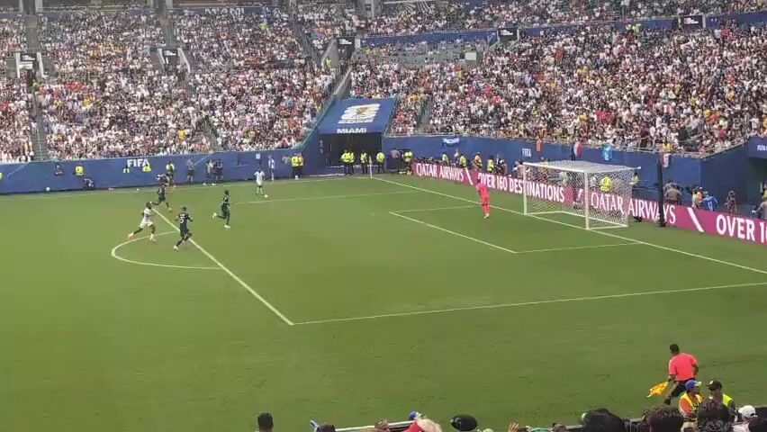 A wide shot shows a soccer game in progress. A player in a dark blue uniform is kicking the ball toward the goal. Another player in a white uniform is nearby. The goalkeeper, also in a dark blue uniform, is positioned in front of the goal. The stands are filled with spectators. The field is green with white lines. The overall scene is energetic and lively, with the focus on the action on the field.