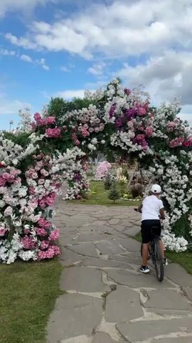 A person on a bicycle rides through a floral archway. The archway is decorated with an abundance of white and pink flowers, as well as green foliage. The path leading to and through the archway is made of large, irregular stone pavers. In the background, beyond the archway, a garden with more flowers and greenery is visible, along with a fountain and some trees. The sky is blue with scattered white clouds. The person on the bicycle is wearing a white t-shirt, black shorts, and a white baseball cap, and they are riding away from the camera. The overall scene suggests a pleasant outdoor setting, possibly a park or a garden.