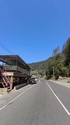 The video shows a clear blue sky above a winding road that leads uphill. On the left side of the road, there are several buildings, including a long structure with a colorful roof and awnings. Some motorcycles are parked in front of these buildings. Further up the road, more buildings and trees can be seen on the hillside. The road itself is paved and marked with white lines. The environment appears to be rural or mountainous, with lush green vegetation on the slopes.