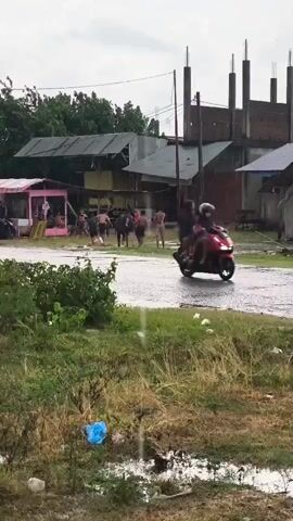 The video shows a group of young people, some shirtless, gathered in an outdoor area. It appears to be raining, with water visible on the ground. In the background, there are several buildings, including what looks like a partially constructed building with exposed concrete pillars, and smaller structures with tin roofs. There are also trees and green vegetation in the foreground and background. The ground is uneven and appears to have puddles of water, suggesting it might be a rural or undeveloped area. The people are scattered around, with some interacting with each other. One person in the center seems to be running. The overall atmosphere is informal and possibly playful.