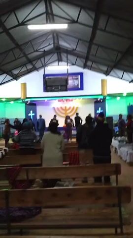 The video shows a group of people in what appears to be a church or a large meeting hall. The people are standing and moving around, some are clapping or raising their hands in the air. There is a stage with a screen and lights at the front of the room. The seating consists of wooden benches. The walls are painted white and purple, and there are blue curtains in the background. The ceiling is corrugated metal with exposed beams. The lighting is provided by overhead fluorescent lights.