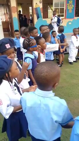 The video captures an outdoor activity involving approximately 20-30 young children, likely of preschool or early elementary age, in Idimu, Nigeria. They are predominantly dressed in blue and white school uniforms, with one child notably wearing a white martial arts-style outfit.

The children are gathered on a green ground cover, appearing to be artificial turf or short grass, and are arranged in loose lines and a circle. An adult woman, presumably a teacher, wearing a red dress, is seen leading the activity, with the children facing towards her, seemingly following instructions. They are actively participating, with audible chanting or singing in unison, which includes numerical sounds.

The setting is an outdoor courtyard or playground of a school, characterized by a light-colored building in the background. The building's walls are decorated with colorful, child-friendly murals, including large pencils, hot air balloons, and a tree with vibrant leaves. The scene is illuminated by bright daylight, suggesting clear weather. The children appear engaged and orderly throughout the activity. No specific text on the building or decorations is clearly legible from the video.