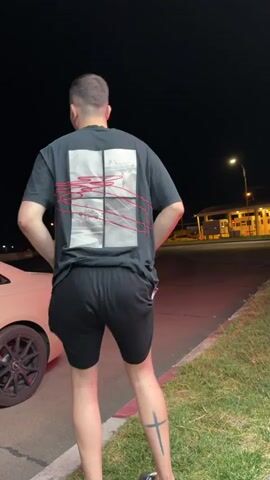 A man in a black t-shirt and black shorts dances outside at night. A light-colored car is parked to his left. In the background, there are streetlights and buildings. The man has a tattoo on his right calf.