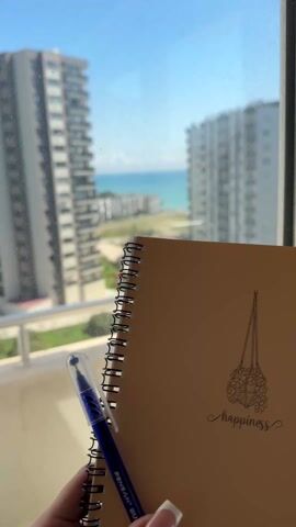 A person holds a spiral-bound notebook with "happiness" written on the cover, next to a blue pen. The background features tall buildings, a beach, and the ocean visible through a window. The sky is blue.