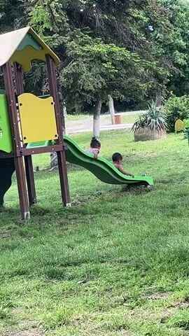 The video shows a children's playground on a grassy field. A green and yellow slide is the main feature. Two young children, one in pink and the other wearing dark clothing, sit on the slide. They aren't moving. There are trees and other greenery around the playground. A trash can sits nearby. The overall scene is outdoors during the daytime.