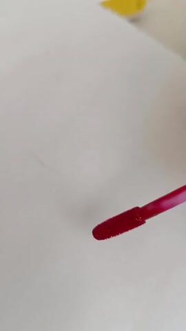 A hand holds a makeup applicator with red liquid and applies it to a white surface. The applicator is moved in a downward motion, leaving a streak of red color. The red liquid drips slightly at the bottom of the streak.