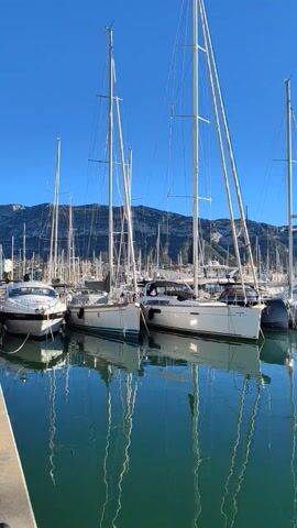 The video shows a harbor on a clear, sunny day. Several sailboats and motorboats are docked, with their masts extending high into the bright blue sky. The water in the harbor is calm and reflects the boats and masts. In the background, a mountainous landscape is visible. The camera pans from left to right, showcasing the variety of boats and the serene environment.
