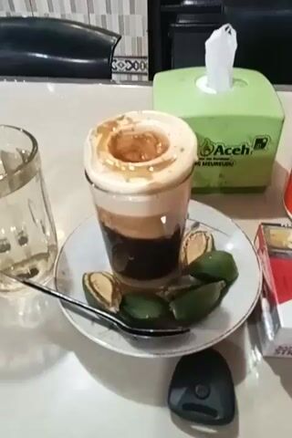 The video shows a table with a coffee drink, a glass of water, a pack of cigarettes, an ashtray, a box of tissues, a few sliced green fruits, a spoon, and a small black object. The coffee drink is in a clear glass on a saucer, and has three distinct layers: a dark bottom layer, a light brown middle layer, and a white foamy top layer with a swirl of darker brown on top. Around the base of the glass on the saucer are several slices of a green fruit. To the right of the saucer is a red and white pack of Marlboro cigarettes and a small orange ashtray. Behind the coffee is a green box of tissues. To the left of the saucer is a glass of water and a spoon. A small black object is just below the saucer. The table has a light-colored, possibly marble or granite, surface. A dark chair is visible in the background.