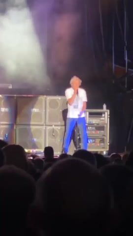 The video shows a male singer with blonde hair, wearing a white t-shirt and blue pants, performing on a stage in front of a large crowd. He is holding a microphone and appears to be singing. Behind him are several large speaker cabinets and audio equipment. There is smoke or haze on stage, illuminated by stage lighting. The perspective is from the audience, looking up at the performer.