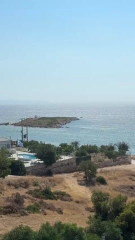 The video shows a pan across a coastal landscape on a clear day. The foreground features dry, grassy terrain with scattered trees and bushes, and a stone wall. A swimming pool is visible to the left. Further back, a road winds along the coastline, with a white car driving on it. The sea is a vibrant turquoise, and a small, rocky island is visible in the distance. The horizon is clear, with a bright blue sky. As the camera pans further, a larger body of water is revealed, bordered by green, tree-covered hills that ascend into mountains. Some buildings are scattered on the hillsides. Towards the end of the video, a resort-like area with lounge chairs and umbrellas appears along the water's edge.