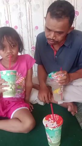 In this media file, a young girl in a pink t-shirt and shorts sits on the floor, holding a cup with a red straw and drinking from it. To her right, a man in a dark blue shirt and light-colored pants is also seated, holding a similar cup with a straw. Another cup with a red lid and straw is visible in the foreground on a green surface. The background features a wall with a patterned wallpaper.

The activity appears to be a casual moment of enjoying drinks, possibly a snack or treat. The girl seems to be engaged in drinking, while the man is looking towards her and appears to be talking or interacting. The man gives a thumbs up gesture at one point. The overall atmosphere seems relaxed and informal.

There is no specific indication of the time of day or weather. The setting is indoors, likely a home environment given the wallpaper and casual seating arrangement. The emotions conveyed are subtle; the girl has a slightly pensive expression, while the man seems engaged and perhaps encouraging.

Visible text on the cups includes "Milo" in white text on a green background on the girl's cup, and a logo that appears to be "Jollibee" with red and yellow accents on the cup in the foreground. The man's cup also has some text and graphics, but they are less clear. The context provided suggests the location is Sumberjaya, Indonesia.