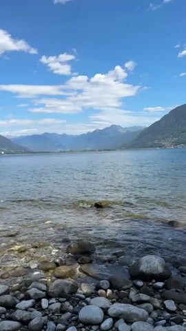 The video shows a view of a lake with mountains in the distance. The foreground is a rocky shore with clear water. The sky is blue with scattered white clouds. The camera is stationary and the video is shot vertically.