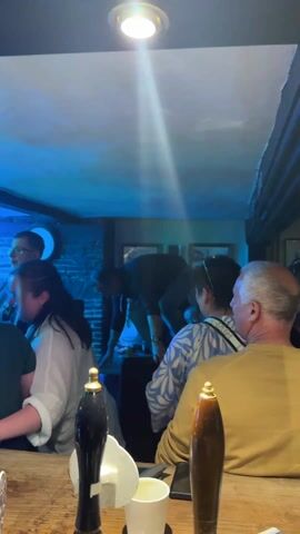 The video shows a woman dancing on the bar counter in a pub.  The pub is crowded with people, some of whom are watching her. There are beer taps on the counter.  The lighting in the pub changes from green to red to blue.  A band is playing in the background.  The woman appears to be having a good time.