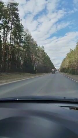 The video is filmed from a first-person perspective inside a moving car, looking forward through the windshield. The car's dark dashboard is visible across the bottom of the frame.

The scene depicts a straight, two-lane asphalt road extending into the distance. Dense forests of tall trees, primarily conifers like pine, line both sides of the road, creating a natural tunnel effect. Some deciduous trees with bare branches are also visible, particularly on the right, suggesting early spring or late autumn. The sky overhead is bright blue with scattered white cumulus clouds.

At the beginning of the video (0:00), a large, multi-axle dump truck, appearing rusty or mud-splattered, approaches from the opposite direction in the left lane. It passes the camera at approximately 0:01. Other smaller vehicles are visible as distant specks on the road, both ahead and behind where the truck was.

The road appears to be in good condition, with a solid white line dividing the two directions of traffic. The weather is clear and sunny, indicated by the bright sky and shadows. No specific text is visible on road signs or vehicles. The setting is a rural highway through a forested area near Solnechnyi, Russia. The overall impression is one of a calm, daytime drive.