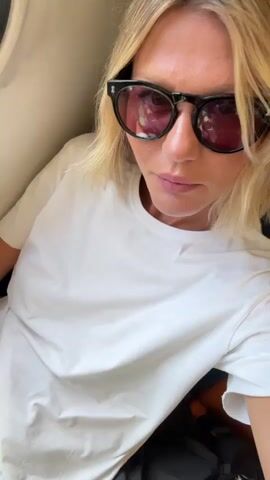 A woman with blonde hair and sunglasses is sitting inside a vehicle, possibly a train. She is wearing a white t-shirt. She is looking downwards. Through the window, a train station platform with a white railing and green trees can be seen. The platform appears to be elevated.