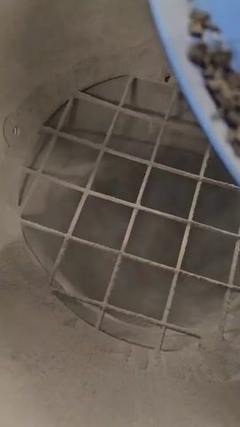 The video shows a close-up, downward-facing view inside a concrete or cement mixer. A blue scoop, partially filled with a gritty, dark material, hovers over the opening. The inside of the mixer is coated with a fine, dusty substance, suggesting it has been used for mixing concrete or similar materials. A metal grate with a grid pattern covers the lower opening of the mixer. The grate appears to be a safety feature or a separator. The overall scene is industrial and utilitarian. The lighting is not clearly indicative of a specific time of day, but the dust suggests an active work environment. No people are visible, but the presence of the scoop implies human operation. The setting is likely a construction site or a concrete production facility in Nowa Wieś Książęca, Poland, given the location context. There is no visible text in the video.