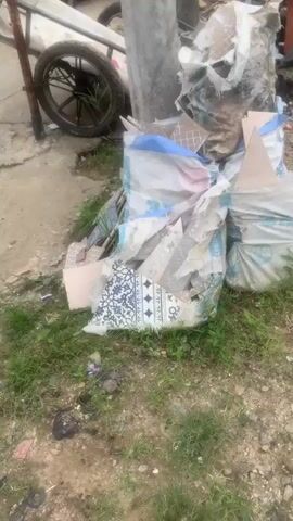 The video shows a pile of construction debris on the ground, including torn bags of cement, broken tiles, rocks, and other materials. The debris is located outdoors, with some grass and dirt visible. In the background, there is a wheelbarrow and some wooden planks. The lighting suggests it is daytime.