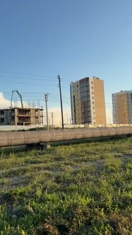 The video shows a panning shot of an outdoor scene with construction activity. In the foreground, a large, insulated pipe runs horizontally across the frame, supported by metal structures. Beyond the pipe, there is green, grassy terrain with some scattered vegetation. In the background, several multi-story buildings are visible. One building appears to be under construction, with visible rebar and incomplete floors. A crane is also present near this building, suggesting ongoing construction work. Other completed or near-completed apartment buildings with windows and balconies are also in view. Power lines and utility poles are present between the foreground and the buildings. The sky is clear and blue, indicating daylight, possibly late afternoon or early evening given the lighting. There are no people directly visible in the shot, but the presence of construction equipment implies human activity related to the building projects.