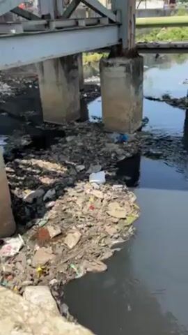 The video shows a body of water heavily polluted with trash. The water itself is dark, almost black, and its surface is covered with large piles of various kinds of debris, including plastic, paper, and other unidentifiable waste materials. The water flows beneath a bridge supported by concrete pillars. The piles of trash are clustered around the pillars and along the edges of the water. In the background, one can see buildings. The video seems to focus on capturing the extent of the pollution issue in this environment.
