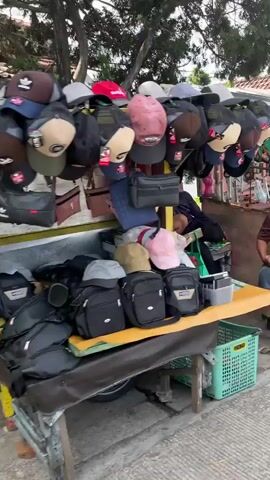 The video shows an outdoor stall selling merchandise. The stall is set up on a wooden table with a yellow top and a metal frame. On display are numerous caps hanging from a rack above the table, and several bags are arranged on the table and in front of it. The caps are of various colors and designs, including some with prominent logos. The bags are mostly black and appear to be crossbody or shoulder bags, with some smaller pouches also visible. A person, wearing a dark top and possibly a headscarf, is partially visible behind the stall, appearing to be attending to the merchandise or resting. The environment appears to be an outdoor setting, possibly a street or market, with trees providing shade overhead. The ground is paved. A green plastic crate with writing on it is visible to the right of the stall. The overall impression is of a small, informal retail operation.