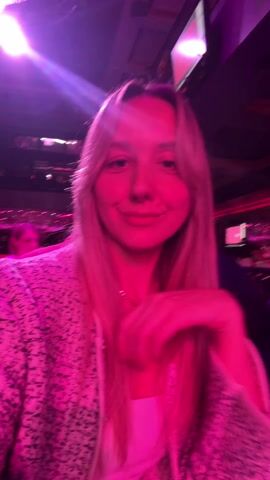 The video shows a woman indoors, likely in a nightclub or entertainment venue, judging by the lighting and the presence of other people in the background. The woman is facing the camera and smiling, with her hand near her face in some shots. She is wearing a light-colored jacket with a subtle pattern, and she appears to have a necklace on. The lighting in the room shifts between pink, purple, and blue, creating a dynamic and vibrant atmosphere. In the background, there are other people, some of whom are walking around, and there appear to be screens or monitors displaying information or visuals overhead. The background activity and the changing lights suggest a lively social setting.