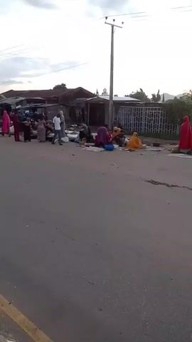 The video shows a street market scene. Several people are present, some dressed in colorful clothing and headscarves, appearing to be vendors and customers. They are gathered along the side of a paved road, with various goods laid out on the ground. Objects visible include bags, baskets, and what appear to be produce or textiles. A few vehicles are also present, including what looks like a red cart or trailer, and a motorized tricycle or tuk-tuk in the distance. Buildings and structures line the opposite side of the road, and a utility pole with wires is visible. The sky is cloudy, suggesting either early morning or late afternoon. The overall impression is of an active outdoor marketplace.