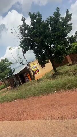 The media file shows a handheld, shaky video of an outdoor scene in Jakatai, Nigeria. The setting appears to be a rural or semi-rural area with dirt roads and some vegetation. Several buildings are visible, ranging from dilapidated structures with corrugated iron roofs to more established brick buildings with red roofs. Some buildings seem to be under construction. In the foreground, a reddish-brown dirt road takes up a significant portion of the frame. Green grass and weeds grow along the side of the road and around the buildings. 

The sky is cloudy with dramatic, puffy white clouds against a blue backdrop, suggesting it might be daytime, possibly late morning or early afternoon. There are no people visible in the video, but a bicycle is leaning against one of the buildings. The overall impression is one of a developing area with a mix of old and new structures, some in disrepair. The movement of the camera suggests the footage was likely taken from a moving vehicle. There are no discernible emotions conveyed, and no clear activity is taking place beyond the general scene of passing through the area. A sign is partially visible on a wall in one frame, but the text is not legible. Overhead power lines are also visible in the upper portion of the frame in some shots.