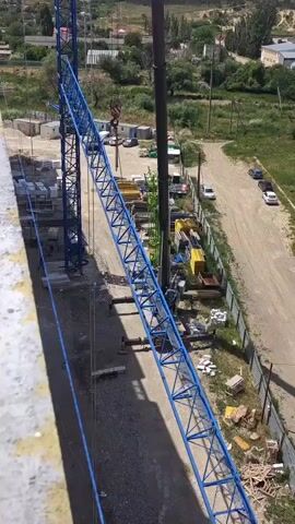 The video shows a construction site from an elevated perspective, possibly from a building under construction. A large blue tower crane is the central focus. Its jib extends diagonally downwards and to the right. A hook is visible hanging from the end of the jib. In the background, there are other construction materials, temporary buildings, a tall smokestack-like structure, and some trees and vegetation. A dirt road runs along the right side of the frame, with a few parked cars visible. The overall scene depicts an active construction environment.