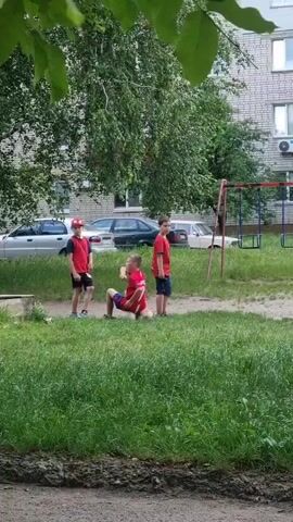 Three young boys are in a grassy courtyard in front of a brick apartment building. The boy in the center is crouched down wearing a red shirt with a white number 7 on the back. The other two boys are standing. One boy on the left is wearing a red shirt and a red baseball cap, and the boy on the right is wearing a red t-shirt and dark shorts. The boy in the center appears to be holding a red disc or frisbee. The boy on the right is pointing his finger towards the center. Cars are parked in the background and a swingset is visible to the right of the boys.