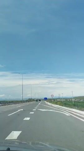 The video shows a view from the perspective of someone driving on a highway on a clear day. The road ahead is paved, with white lane markings and directional arrows. There are streetlights on the left side of the road. In the distance, there are a few cars and a blue highway sign. On the right side of the road, there is a speed limit sign indicating 90. Beyond the road, there are fields and hills under a blue sky with scattered clouds.