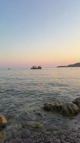 A red boat with people on it is sailing on the sea during sunset. The sky is painted with soft pink and orange hues, transitioning into a clear blue. The water is calm with gentle waves. Rocks and pebbles are visible in the foreground on the shore. Some people are standing on the boat, appearing to be enjoying the scenery. The overall atmosphere is serene and peaceful.