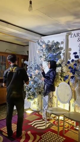 A wedding photoshoot is taking place in a room decorated with white and blue floral arrangements. A person wearing a dark blue kebayah and a black hijab poses for a photo, holding a flower. The background features white drapery and a white backdrop with floral embellishments. A person in a black t-shirt with a "W INHU WEDDING" logo is directing the photoshoot. The room is lit by artificial light. Two white chairs with woven seats are visible. The floor is covered with a patterned rug. The time appears to be evening or indoors with artificial lighting. The overall mood is celebratory and formal, typical of a wedding event.