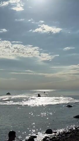 The video captures a serene waterscape under a bright, partially cloudy sky. The sun, high in the sky, casts a brilliant, shimmering reflection across the water, creating a sparkling path.

In the mid-ground, two individuals are riding jet skis on the calm water. One jet skier is actively maneuvering, creating white foamy wakes as they turn, while the other appears to be cruising further in the distance. Their silhouettes are stark against the sunlit water.

In the foreground, the rocky shoreline is visible, with dark, smooth stones lining the edge of the water. Several people are sitting or standing on the shore, their figures also silhouetted against the bright background, seemingly watching the activity on the water. The overall impression is one of leisure and outdoor recreation in a coastal setting.