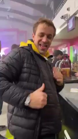 The video begins with a smiling man in a black puffer jacket and a yellow hoodie giving a thumbs up in front of a bar. He has a wristwatch. The camera then focuses on a bar counter with multiple plastic cups filled with beer. The cups feature an image of a man with a mustache and the words "JACQUES FROMAGE." The cups are decorated with images of cheese. A hand picks up one of the cups. The bar has a red, green, and white color scheme. People are visible in the background, and there is a sign that reads "BERNAD." The lighting is bright, and the environment appears festive, possibly a party or event.
