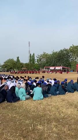 The video shows a wide shot of a grassy field with a large group of people sitting on the ground. The people are facing a stage or platform in the distance, where there appears to be an event or performance taking place. Many of the people are wearing uniforms, some of which are blue and others are teal or light green. Some are wearing headscarves, indicating they are likely students. The background features trees, a tall communication tower, and a white fence with a goalpost. The sky is overcast. The camera is positioned behind the seated audience, looking towards the distant activity.