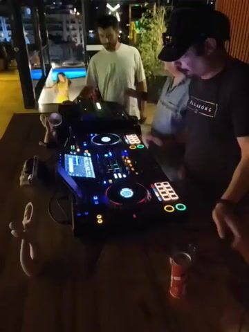A group of three men stand behind a DJ booth at night. The DJ booth features a professional DJ setup with multiple screens, turntables, and mixing boards. The lighting is dim and atmospheric, with colored lights illuminating the equipment. The men appear to be engaged in conversation or operating the DJ equipment. In the background, a swimming pool and distant city lights are visible through a large window. The overall scene suggests a social gathering or event taking place at a venue with a rooftop or elevated view.