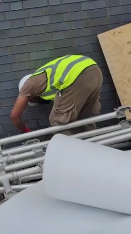 The image shows a construction worker from the back, wearing a white hard hat, a brown t-shirt, khaki work pants with large cargo pockets, and a bright yellow safety vest with reflective stripes. They are also wearing red gloves. The worker is bent over, appearing to be engaged in some task near a grey brick wall. In the foreground, there are stacks of metal scaffolding poles and large white cylindrical objects, possibly pipework or insulation. To the right of the worker, a piece of light-colored plywood is leaning against the brick wall. The scene suggests an outdoor construction site, likely during daylight hours, given the lighting. No text is visible in the image.