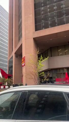 The video shows a street corner on a rainy day. A tall building of pink-brown stone is visible, with a sign for Muf Med Bistro above its entrance. A modern sculpture of a teardrop with red horizontal platforms inside stands prominently in front of the building. The sculpture sits on a brick base, surrounded by small plants and a mirrored sphere. A person in dark clothing walks past the building. A white car is partially visible at the bottom of the frame in the initial seconds. A traffic light is visible. The street is wet from the rain, reflecting the surroundings.