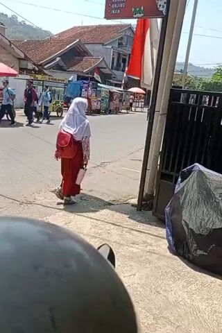 A person wearing a white hijab and red skirt walks away from the camera on a street. They are carrying a pink water bottle in their right hand and a red backpack on their back. Several food carts are visible on the left side of the street, with people around them. A white building with a red roof stands behind the food carts. In the background, a hillside is visible. To the right of the frame, a black metal fence and a large black tarp-covered object are present. A motorcycle helmet is partially visible in the foreground, suggesting the video is filmed from the perspective of someone on a motorcycle. Another person stands on the right side of the street, looking at their phone, as a motorcycle approaches.