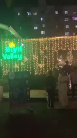 At night, the entrance to the "Biryd Valley" establishment, adorned with string lights and lush greenery, is bustling with people. A brightly lit sign displays "Happy Hours," while a multi-story building serves as a backdrop. Individuals are seen entering and exiting the establishment, some dressed in formal attire. The scene exudes a festive ambiance with a hint of elegance.