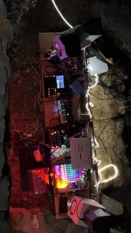 The video shows a collection of electronic music equipment set up outdoors at night. The equipment is arranged on a long, narrow table or surface, illuminated by string lights and the colorful lights from the devices themselves. The background appears to be a rocky or natural environment. The devices include synthesizers, drum machines, mixers, and possibly speakers. One device has a screen displaying text or a waveform. There are numerous cables connecting the various components. The camera angle is overhead and the video is rotated 90 degrees counterclockwise.