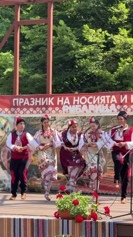A group of five young people, three females and two males, are dancing on an outdoor stage. They are wearing traditional Bulgarian folk costumes. The males wear black pants, white shirts, and red vests embroidered with black and white designs. The females wear long, embroidered skirts and blouses, with vests and floral headdresses. They are holding hands in a circle and performing a folk dance. Behind them is a banner with red and white floral patterns and Cyrillic text. The stage is set against a backdrop of lush green trees. Red geraniums in pots are placed on the edge of the stage in front of the dancers.