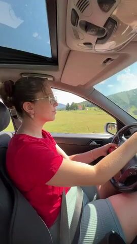 A woman wearing a red t-shirt and glasses is driving a car with a sunroof open. She is wearing a seatbelt and is looking forward. The car is moving along a road through a rural landscape with green hills and trees. The sky is blue with scattered white clouds. The interior of the car is visible, including the dashboard, steering wheel, and a radio.