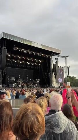 A concert stage at the "Summer Sessions" festival is visible with a large crowd of people in the foreground. The stage is equipped with lighting rigs and screens, and a band appears to be performing or setting up. The audience is diverse in age and gender, with many people holding up their phones or drinks. The sky is overcast.
