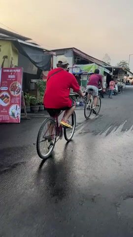 The video shows two individuals riding bicycles down a street lined with buildings. The person in the foreground, facing away from the camera, is wearing a red t-shirt, shorts, and a baseball cap. They are riding a red bicycle. Behind them, another person, also facing away, is wearing a maroon t-shirt and white shorts. This person is riding a white bicycle. Several motorcycles are parked along the side of the street. The environment appears to be a commercial area with small shops or stalls. The time of day seems to be late afternoon or early evening, given the lighting conditions.
